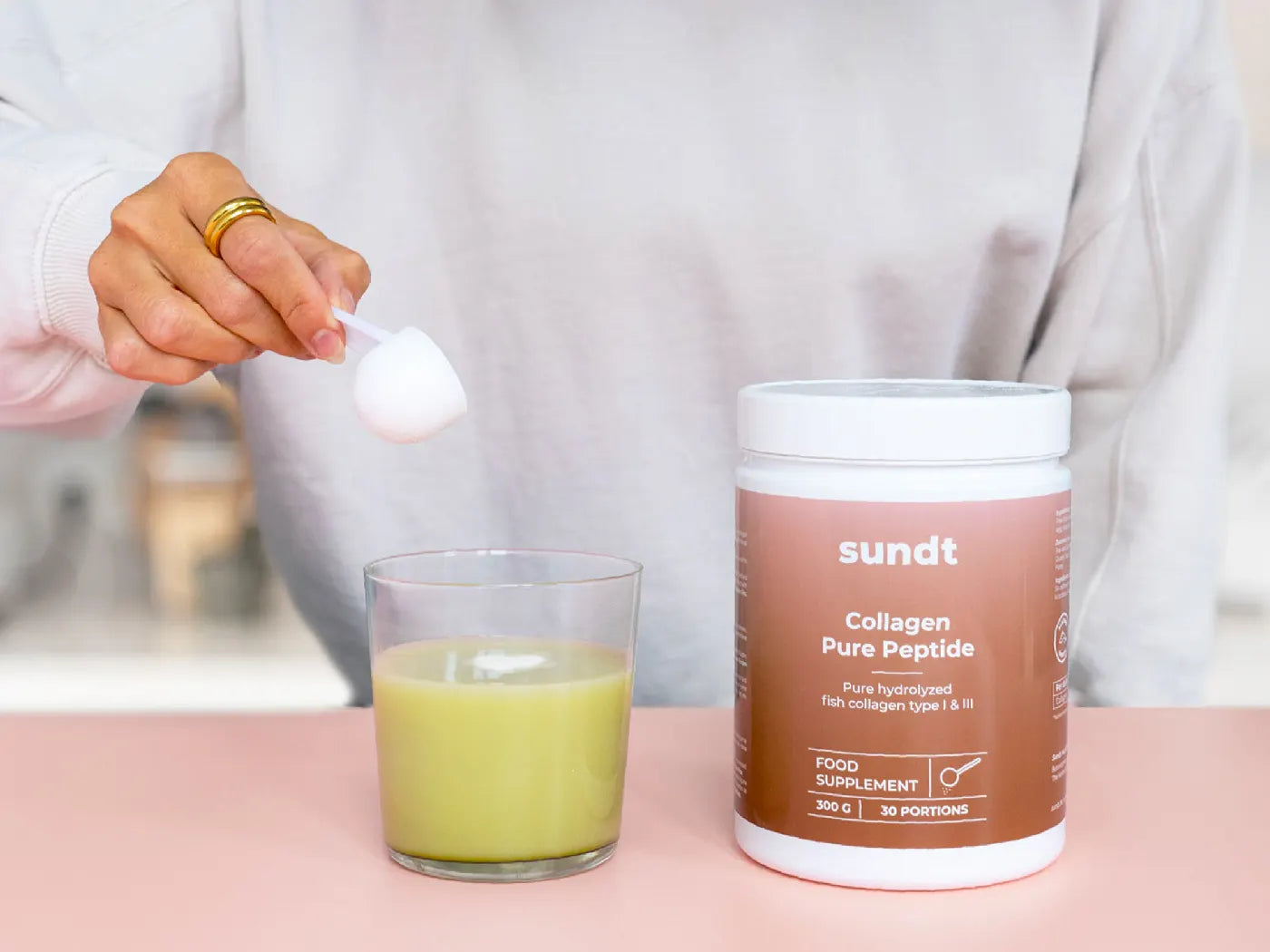 Collagen Produkt von Sundt, das von einer Hand zubereitet wird. Es wird auf einer rosafarbenen Unterlage präsentiert.