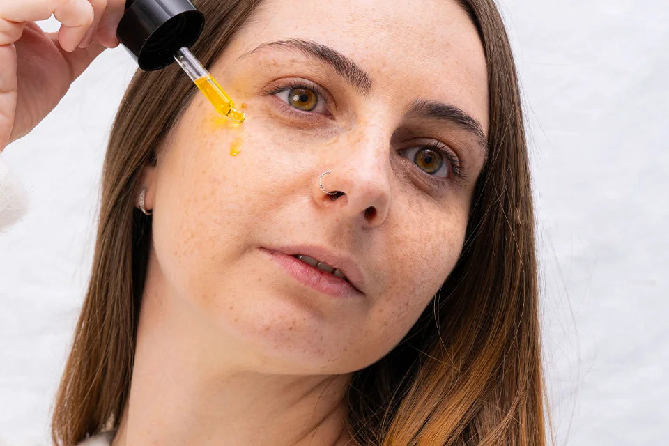 Eine braunhaarige Frau träufelt mit einer Pipette Gesichtsöl auf ihre Haut.