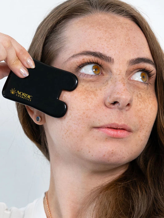Eine Frau mit braunen Haaren hält sich Obsidian Gua Sha von Nordic Cosmetics vor die Wangen.