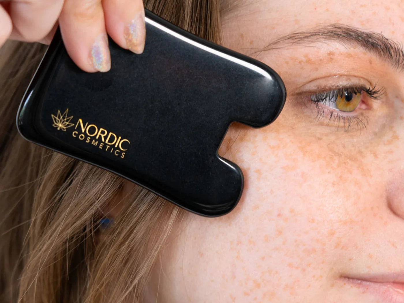 Eine Frau mit braunen Haaren hält sich Obsidian Gua Sha von Nordic Cosmetics neben ihren Augen