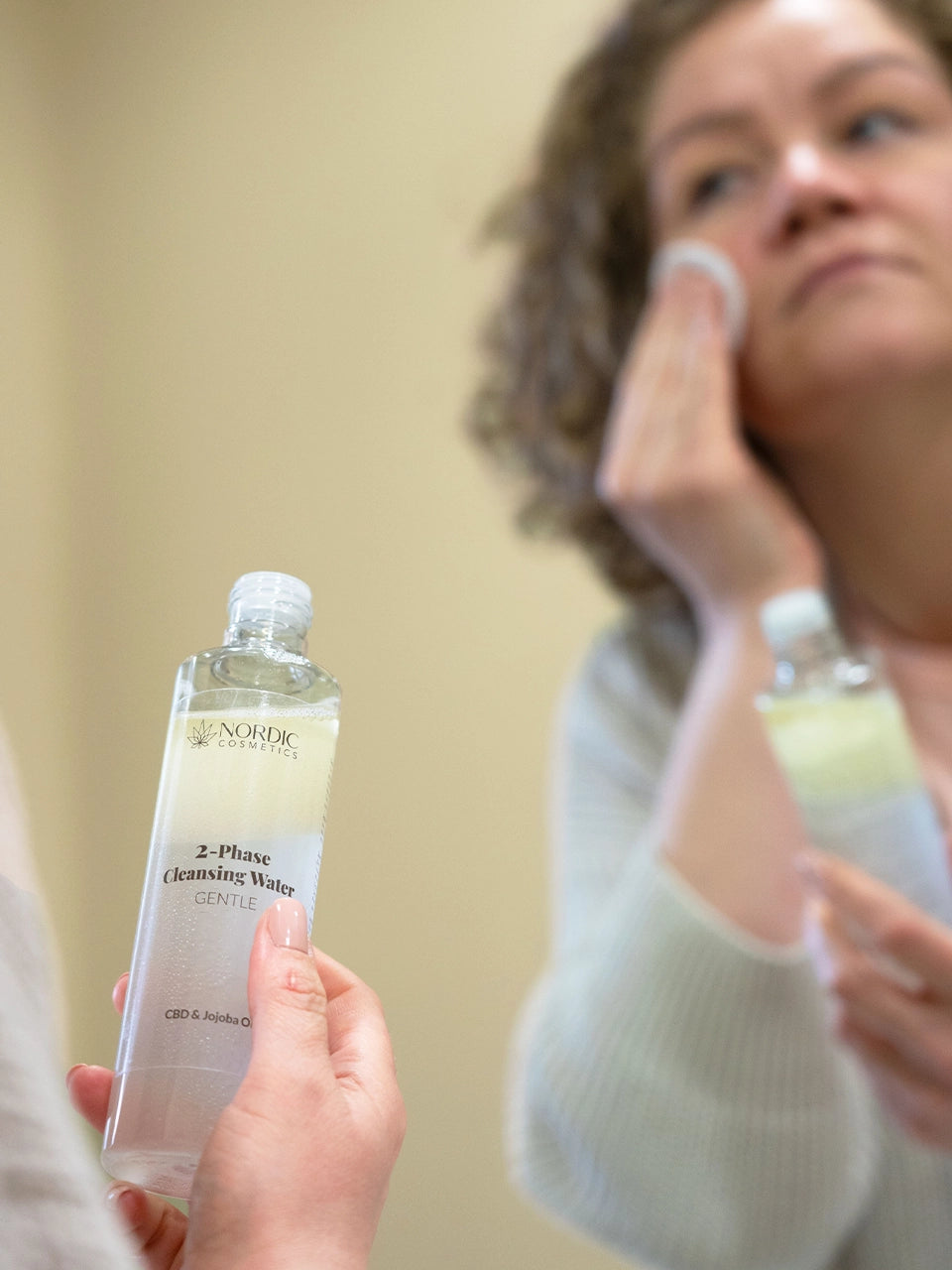 Die Frau benutzt 2-Phase Cleansing Wasser mit CBD von Nordic Cosmetics. Sie tupft das Produkt mit einem Wattepad über ihr Gesicht. Sie steht vor dem Spiegel.